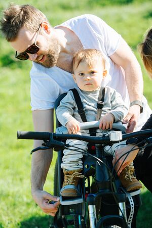 Attentive father carefully strapping his kid to front bike baby chair. Over green countryside. Child looking at camera, he is not very fond of process.の写真素材