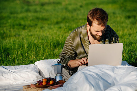 Man sitting in bed outdoors in grassfield in the middle of nowhere, keen working on his laptop under...
