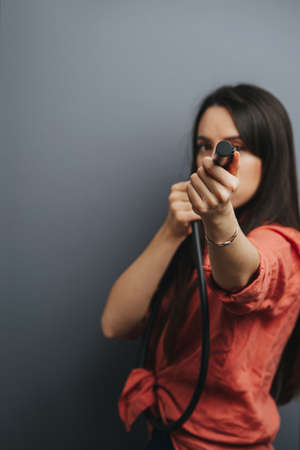 Pretty young woman with a serious face, playing around with a shower nozzle, aiming at you. Blurred, focus on a cealed tip. Over gray background.の写真素材