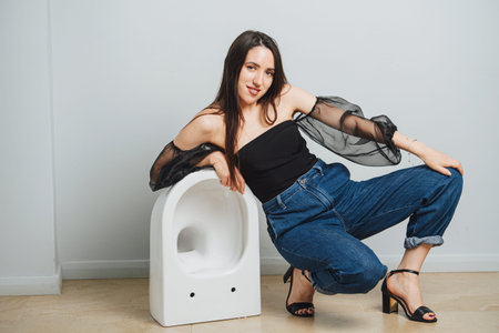 Attractive young woman in fashionable clothes crouching down on one leg, leaning on a toilet bowl....