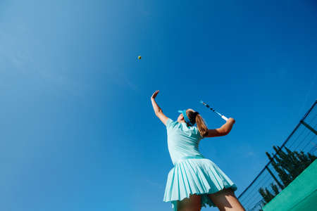 Teenage girl in a sky blue sportive outfit playing tennis, throwing ball up in the airの写真素材