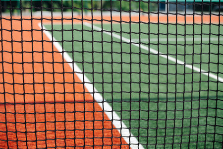 Part of a new empty tennis court captured through a net on a sunny day