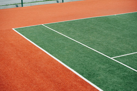 Corner of a green inner tennis court with white markings and brown outer part. Low angle shot.
