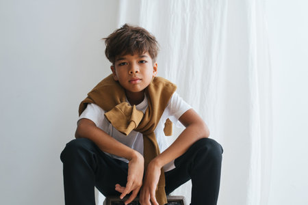 Emotionless lame indian ten year old asian kid looking at camera, sitting low, knees up. Indoors,...