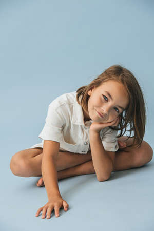 Smiling ten year old little girl in a white thick romper suit sitting on the floor, leaning on her hand, playing cute. Over blue background.の写真素材