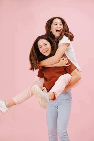 Beautiful little twin sisters happily playing together, one riding the other. Over pink, studio shot. They wear pink and blue pants in contrast with their shirts. Their hair move from motion.の写真素材