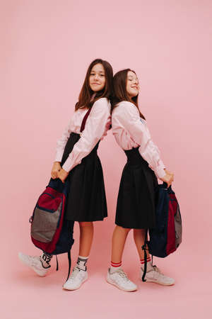 Beautiful little twin schoolgirls happily posing with their backpacks, holding them in hands. Leaning their backs on each other. Low angle. Over pink background, studio shot.の写真素材