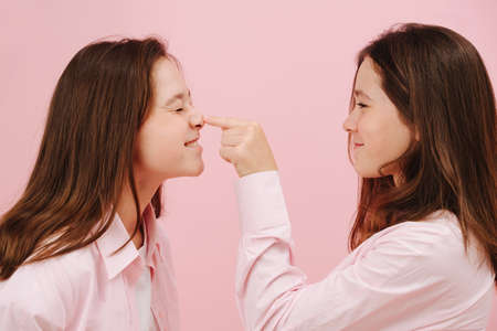 Beautiful little twin sisters happily playing, one pressing on nose of the other. Over pink background, studio shot. One is amused, second is squinting.の写真素材