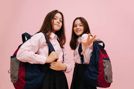Beautiful little twin schoolgirls happily posing with their backpacks, looking at the camera. Low angle. Over pink background, studio shot. One girl extending hand to the cameraの写真素材