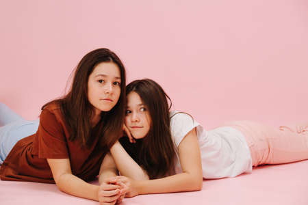 Beautiful little twin sisters happily lying on the floor on their stomachs. Over pink background, studio shot. One looking at camera, second making funny face.の写真素材