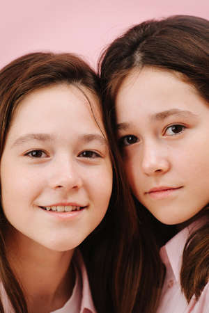 Beautiful little twin sisters pressing heads to get into image frame. Over pink background, studio shot. Both looking at the camera, one smiles openly, her sister has closed smile.の写真素材