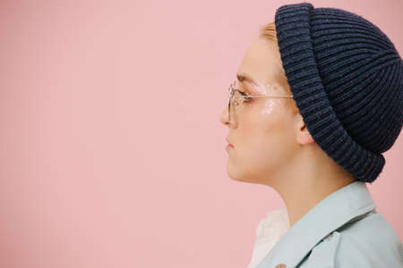 Side portrait of young resolved woman in a cap, round glasses and with serious face. Over pink background. Model has short hair and stars eye make up.の写真素材
