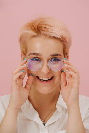 Portrait of a smiling young woman with short dyed blond hair and round glasses and stars eye makeup....