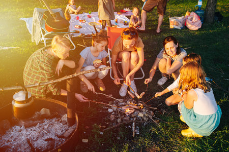 Friends and family gathered around bonfire, having picnic. They are cooking sausages on a bonfire. On a spring sunny dayの写真素材