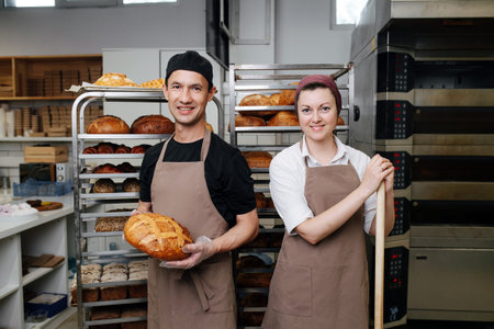 Two bakers, male and female posing for a photo in a bakery kitchen. Over Iron tray shelves filled...