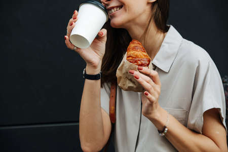Smilng mature woman on the street drinking coffee with wrapped in paper croissant. Close up, cropped face.の写真素材