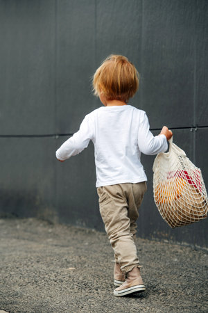 Little boy with blond hair carrying net bag with fresh crispy bread, outdoors on the street, walking...