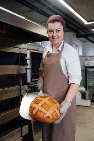 Female baker presnting fresh bread on a special flat shovel. She's wearing headband, smiling.