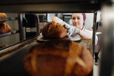 Female baker placing fresh bread on a shelf, using special flat showel. She's wearing headband....