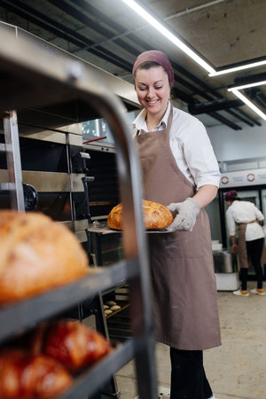 Happy smiling female baker with fresh bread on a flat shovel to place on a shelf. She's wearing...