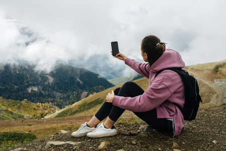 Young woman taking a picture, sitting on the ground high up in mountains covered with short grass and trees, low clouds hanging around her. She's wearing hoodie, yoga pants and backpack.の写真素材