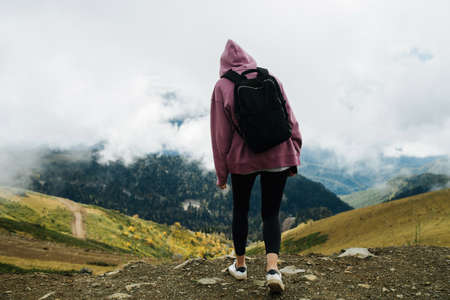 Young woman in mountains covered with short grass and trees, low clouds hanging around her. She's wearing hoodie, yoga pants and backpack, Enjoying the view.の写真素材