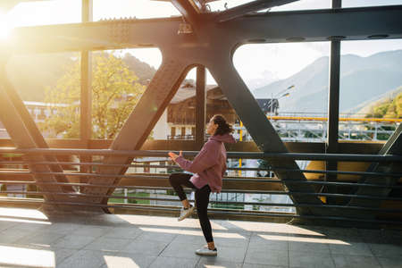 Young woman exercising on a steel girder walking bridge with a view on houses with wooden top parts and mountain forest. She's wearing pink hoodie and yoga pants.の写真素材