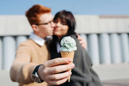 Blurred image of couple sitting outdoors, kissing. Guy holding ice cream in outstretched hand, blurred face. On an autumn sunny day, both wearing sweaters with white shirt collars.の写真素材