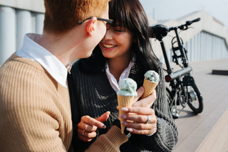 Carefree couple sitting outdoors, crossing hands with ice cream, sensually touching with foreheads...