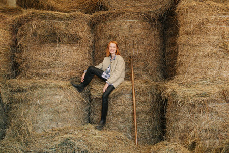 Ginger girl with lovely freckles sitting on piled up hay bundles inside a barn. She wears beige...