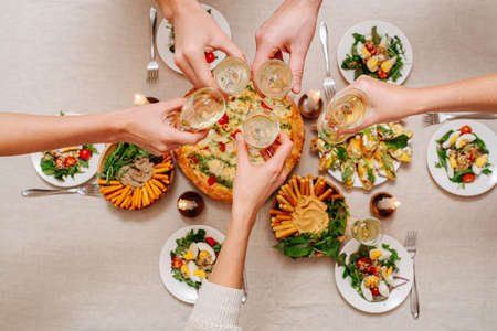 Overhead view of hands clinking glasses over a table with Italian cuisine assorti. Margarita, cheese sticks with hummus and arugula, salad with tomatoes and eggs served with white wine.の写真素材