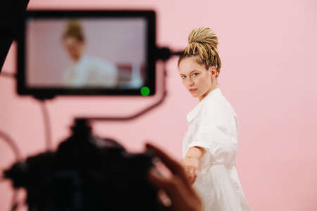 Attractive woman with blond afro braids in a bun posing in front of a camera with a field monito. Over pink background. Blurred equipment in a foreground.の写真素材