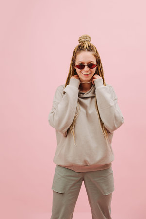 Entertained cool looking young woman in narrow sunglasses with stylish blond afro braids over pink...