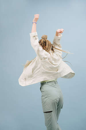 Celebrating young woman with stylish blond afro braids from behind over blue. Over blue background. Throwing hands in the air.の写真素材