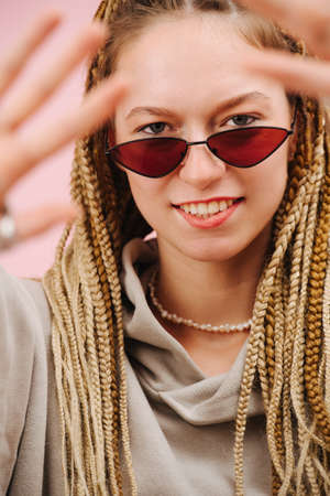 Eccentric smiling young woman in narrow sunglasses with stylish blond afro braids over pink background. Looking at camera over sunglasses, blurred fingers in front of the face.の写真素材