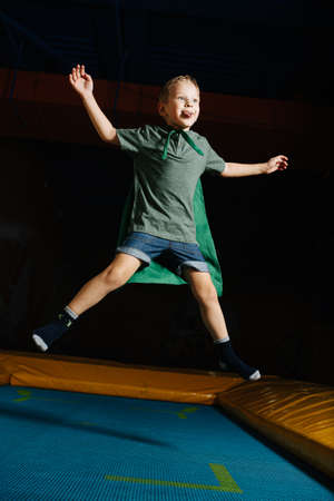 Confident boy in a cape jumping on trampoline in entertainment centerの写真素材