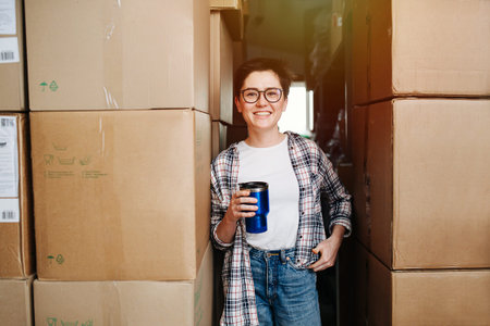 Emotional friendly female warehouse worker standing with coffee between boxes