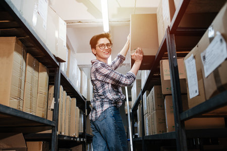 Happy female warehouse worker putting heavy cardboard box on a top shelfの写真素材