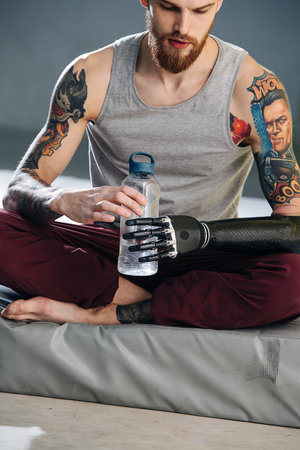 Worn out cyborg man with bionic hand sitting on a mat, opening water bottleの写真素材