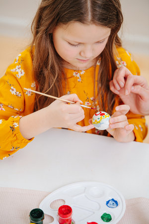 Smiling happy little girl painting easter eggs behind a table using brushの写真素材