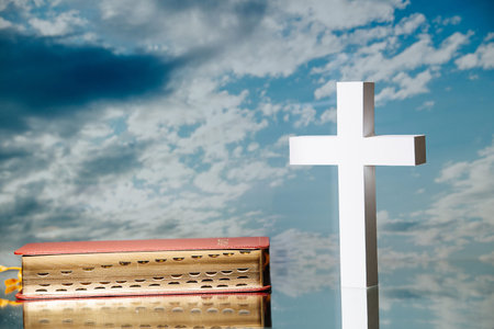 Bright white cross next to a holy bible against blue clouded sky.