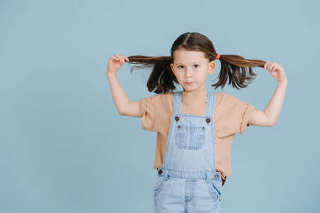 Cute little girl pulling her ponytails to sides, posing for a photo over blue background. She wears jeans overalls and brown shirt.の写真素材
