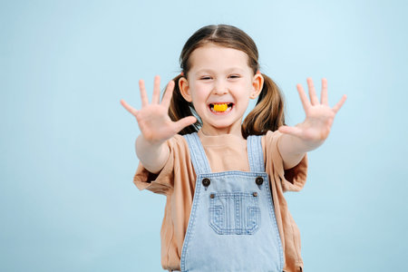 Funny little girl posing with candy in her teeth over blue background. She has two ponytails, wears overalls. Her hands outstretched forward with fingers spread apart.の写真素材