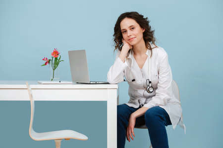 Beautiful young doctor waiting for a patient in her office, looking at the camera, posing for a photo. Over blue background. She wears white robe and stethoscope.の写真素材