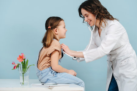Pleasant young female doctor examining happy social little girl with a stethoscope. Side view. Over blue background.の写真素材