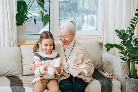 Cozy home environment, grandmother teaches her granddaughter to embroider, they sit on the sofa at...