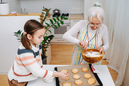 Keen granny sprinkling sugar powder on fresh cookies next to her granddaughter. Spending time...