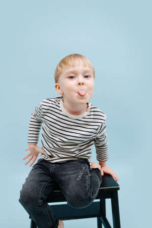 Naughty little blond boy sitting on a stepping stool, stucking his tongue out. He's wearing striped long-sleeve shirt. Studio shot. Over blue background.の写真素材
