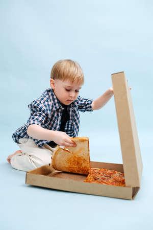 Hungry little blond boy sitting on the floor, tearing pizza slice for himselfの写真素材