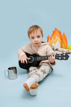 Adorable cute little boy sitting next to a fake campfire with paper flames and birch logs. He is...